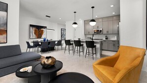 A modern living room with a sofa, a coffee table, and a yellow armchair. There is a dining area with a table and chairs, and a kitchen with cabinets and appliances. - Hazel’s Hideout | Big, PVT retreat near Universal w/ island theme RM, pool, more (Orlando)