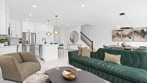 Eine moderne Küche mit weißen Schränken und Edelstahlgeräten, ein grünes Sofa mit Kissen sowie ein runder Couchtisch mit einer Schale und einem Glas.