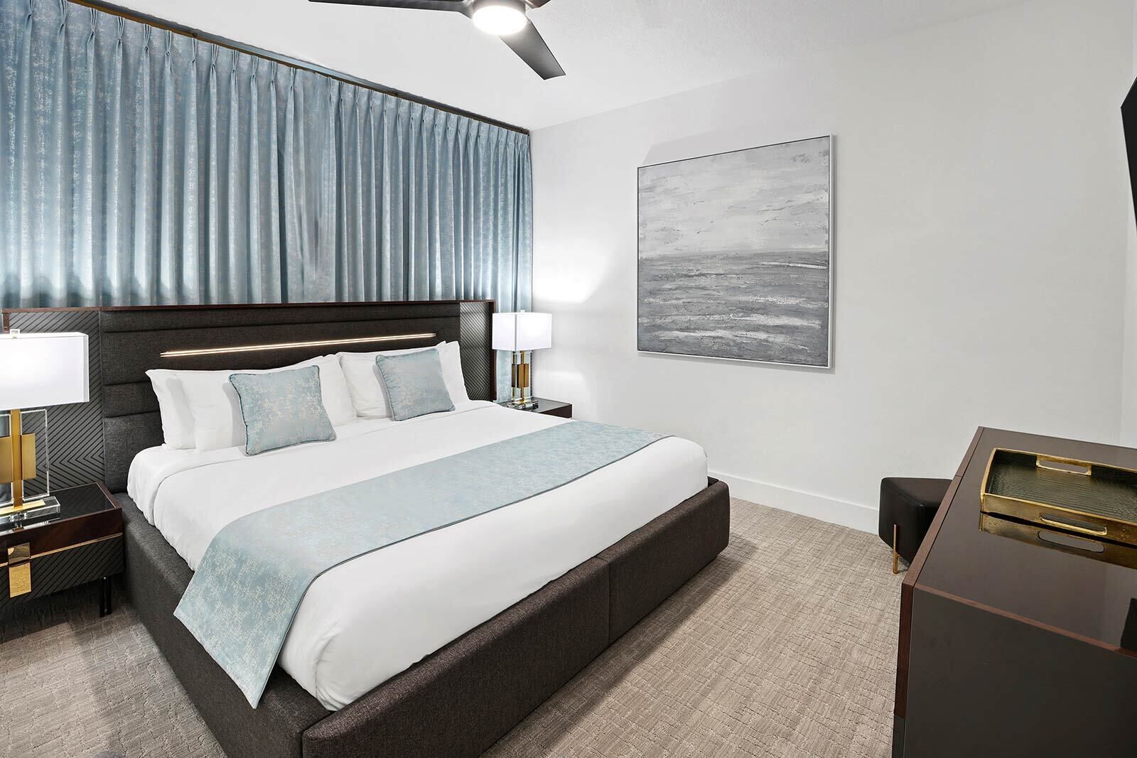 Quarto de hotel moderno com uma cama grande, duas mesas de cabeceira com luminárias, um ventilador de teto e uma janela ampla com cortinas.
