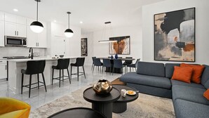 A modern kitchen with white cabinets, a dining area with black chairs, and a living room with a grey sofa and orange cushions. - Rory’s Residence | Instaworthy home near parks w social media BR, pool, luxe BRs (Orlando)