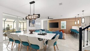 Une cuisine moderne dotée d’un îlot central, un coin repas avec une table blanche et des chaises turquoise, et un salon avec un canapé et un téléviseur.