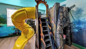 Un espace de jeux avec un toboggan jaune, une échelle et une fresque représentant un volcan.