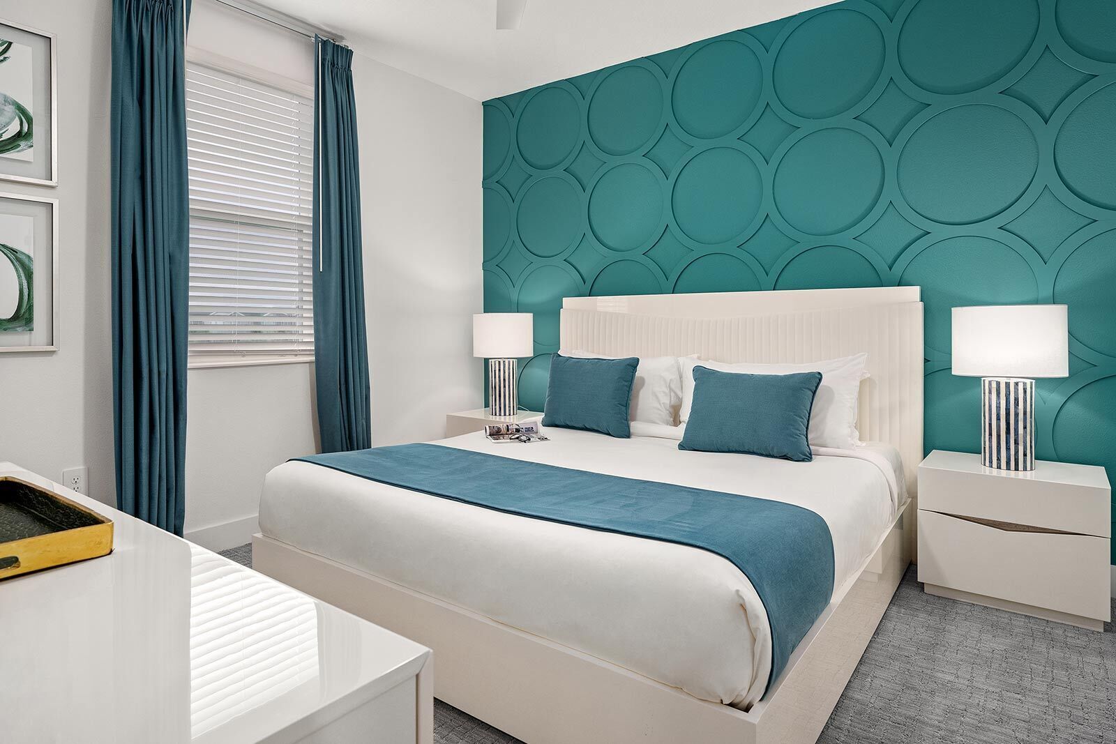 Una habitación de hotel moderna con una pared de acento color verde azulado, una cama blanca con ropa de cama azul y dos mesitas de noche con lámparas.