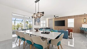Een moderne woonkamer met een grote televisie, een eethoek met een witte tafel en blauwgroene stoelen, en uitzicht op het groen.