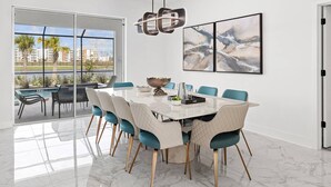 Een moderne eetruimte met een grote tafel, blauw-witte stoelen en uitzicht op een zwembad en gebouwen door grote ramen.