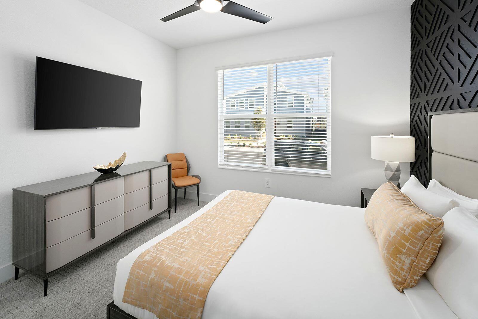 A modern hotel room with a bed, a desk, a chair, a TV, a window with blinds, and a ceiling fan.