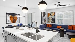 A modern kitchen with a central island, a dining area with a round table, and a living room with a blue sofa and orange cushions.