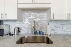 Een keuken met witte kasten, een roestvrijstalen spoelbak, een waterkoker en een betegelde achterwand.