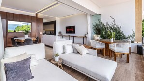A modern living room with a sofa, dining table, and chairs. There is a kitchen area in the background, a TV mounted on the wall, and a balcony with plants. - Largest Rooftop Pool in Latin America! (Puerto Vallarta)