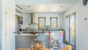 A modern kitchen with a central island, two windows, and a view of the outdoors. - Villa Cycladic View Syros  (Azolimnos Syros)