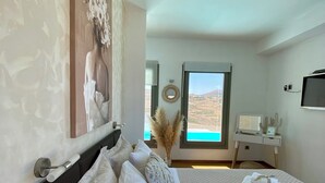 A bedroom with a bed, a painting, a window with a view, a desk, and a television. - Villa Cycladic View Syros  (Azolimnos Syros)