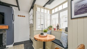 Un rincón acogedor con una mesa redonda de madera, un banco con cojines estampados y vistas a través de contraventanas blancas.