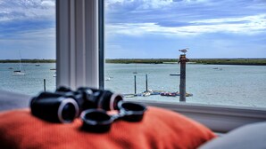 Vista de un cuerpo de agua con barcos, una gaviota sobre un poste, y una ventana con un par de gafas sobre un cojín.