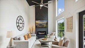 A modern living room with a fireplace, a ceiling fan, and a large window offering a view of the outdoors. - Sunset Chalet 2 Bedroom Cabin by RedAwning (Gatlinburg)