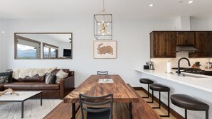 A modern kitchen with wooden cabinets, a central island with bar stools, a dining table with chairs, and a large window with a mountain view. - Hay Meadow Lodge 3 Bedroom Townhouse by RedAwning (Fraser)