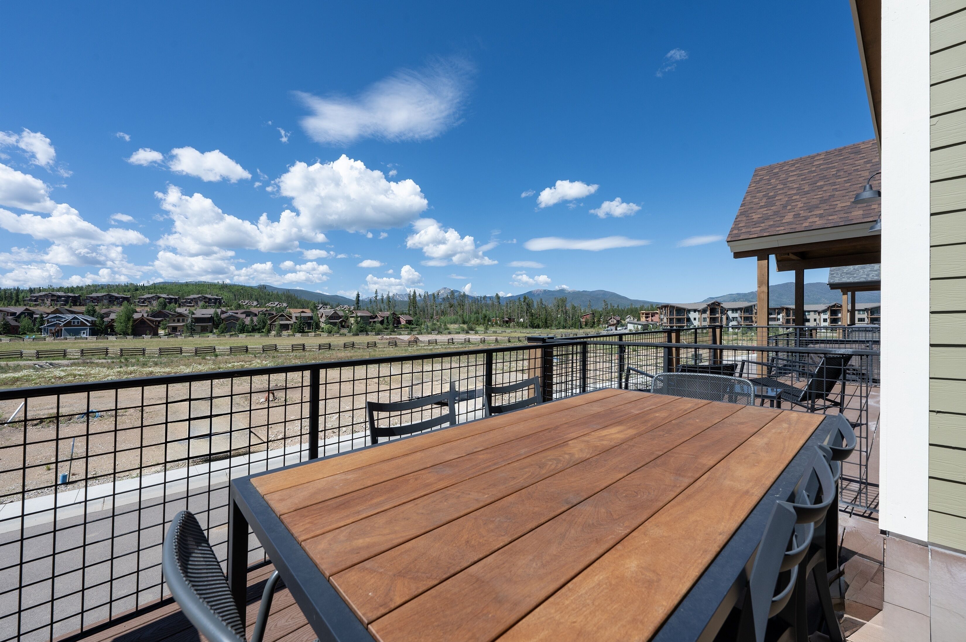 An outdoor patio with a wooden table and chairs, a metal fence, and a view of a residential area with houses and mountains in the background.