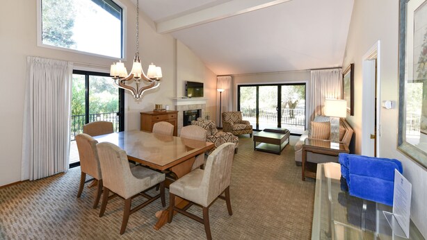 A spacious living room with a large dining table, comfortable seating, a fireplace, and large windows offering a view of the outdoors.