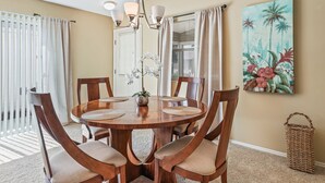 A dining area with a round wooden table, six chairs, a hanging light fixture, and a tropical-themed painting on the wall.