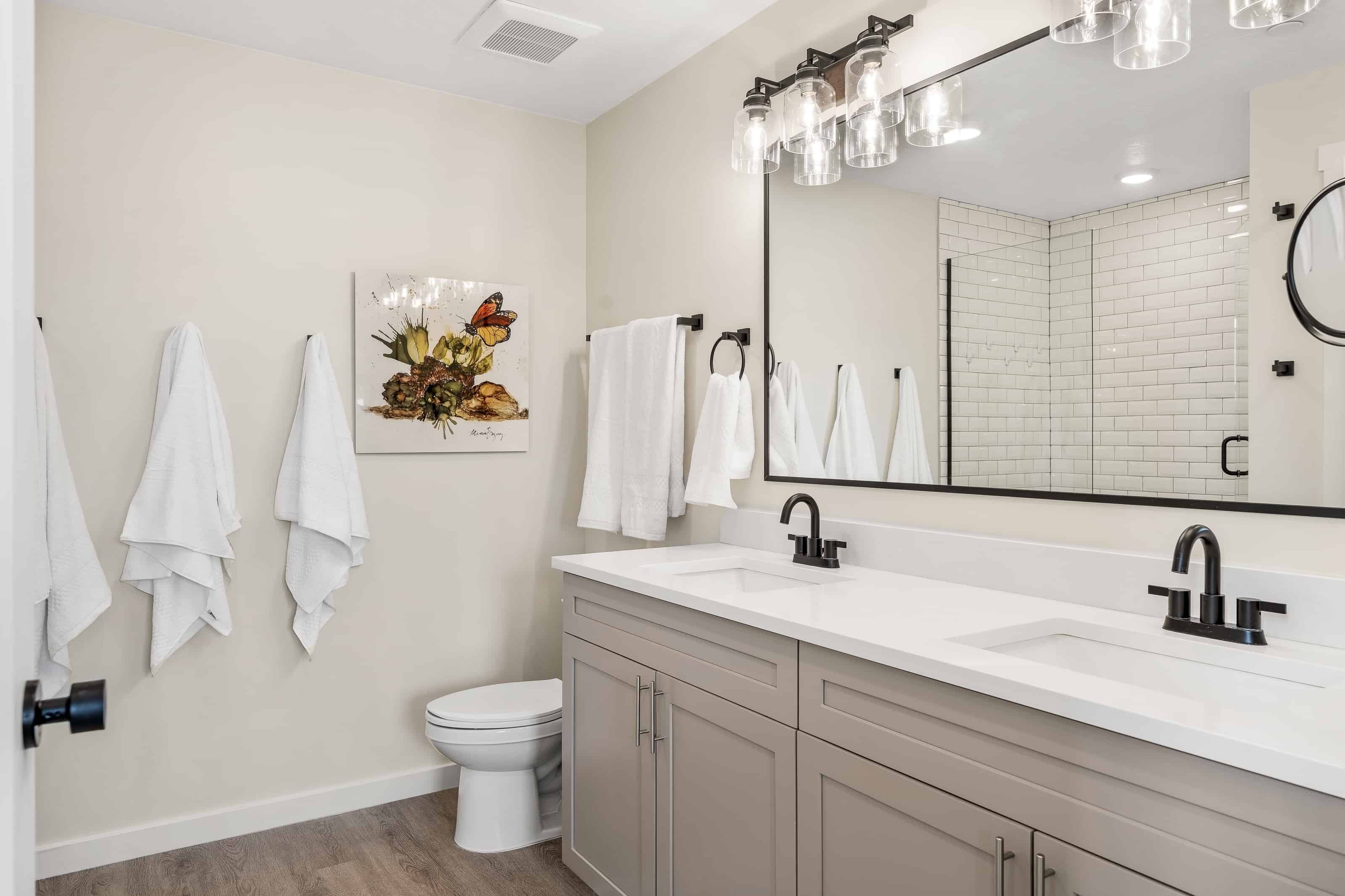 A modern bathroom with a white sink, black faucet, and a large mirror with a light fixture above it. There are white towels hanging on hooks and a toilet visible.