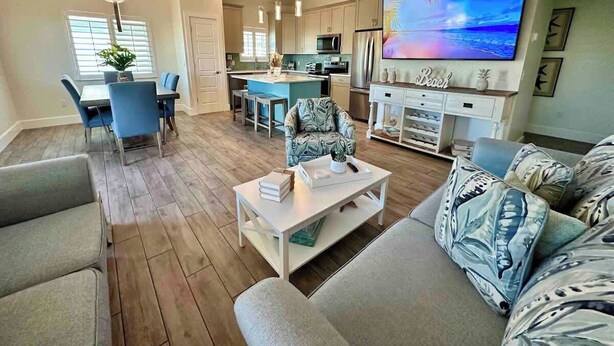 A modern living room with a sofa, coffee table, and TV. There is a dining area with chairs and a table. The kitchen has a large island and modern appliances. - Beyond The Sea · 70+ 5-Star ·  Walk to Beach-Town (Fort Myers Beach)