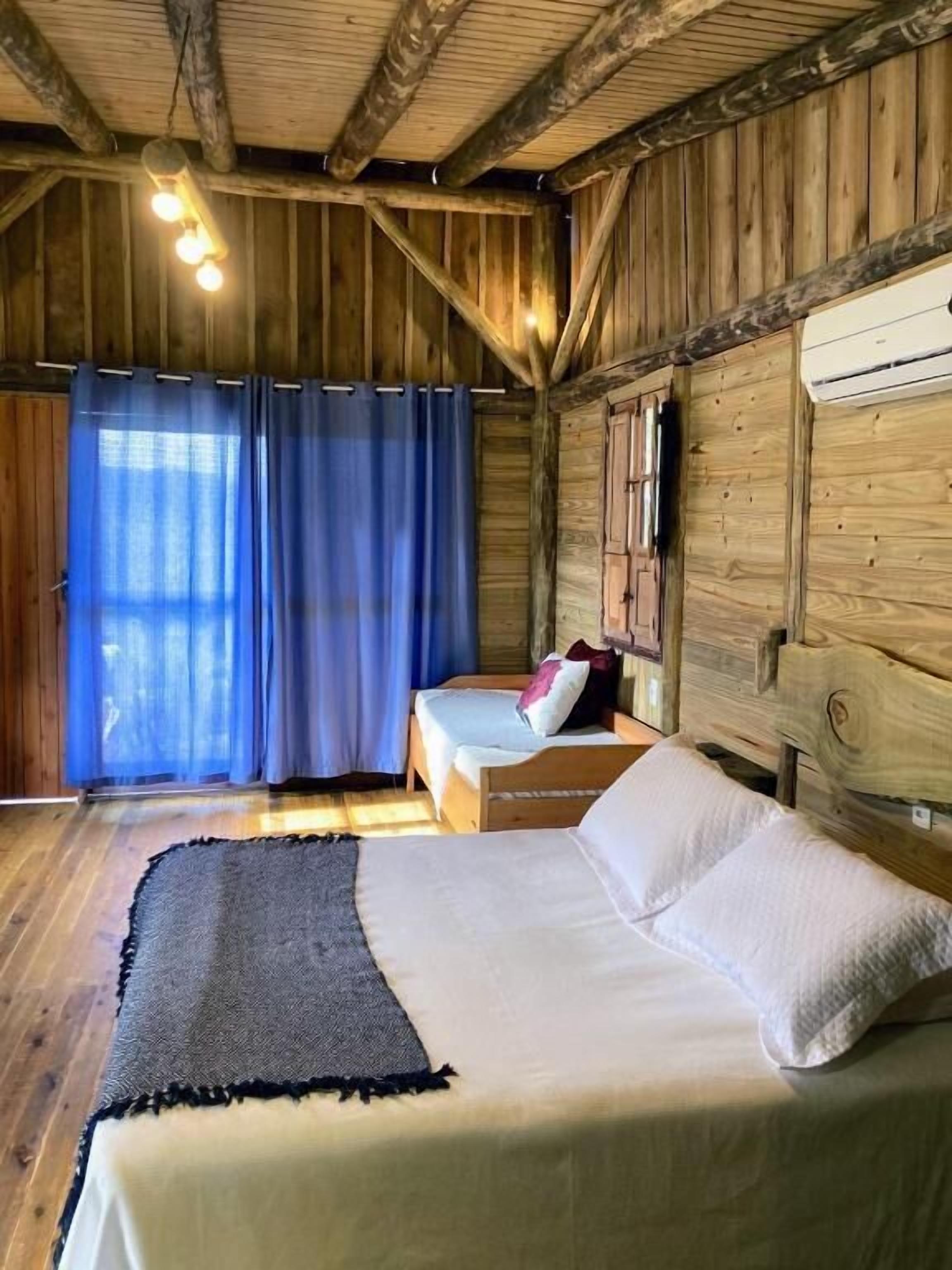 Una habitación de cabaña de madera con una cama, un banco y una ventana con cortinas azules.