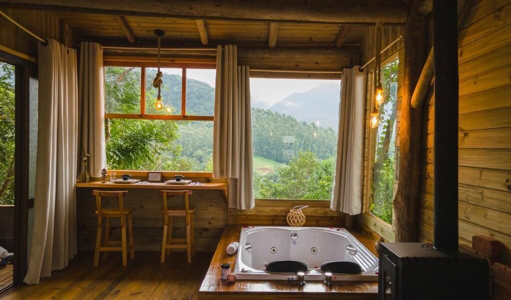 Uma cabana rústica com interior de madeira, uma mesa de madeira com cadeiras, uma janela grande com cortinas e uma lareira a lenha.
