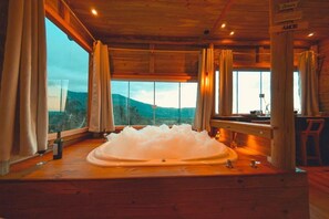 A wooden cabin with a large bathtub in the center, a window with a view of the mountains, and a wooden ceiling. - Pousada Flor De Lótus (Praia Grande)