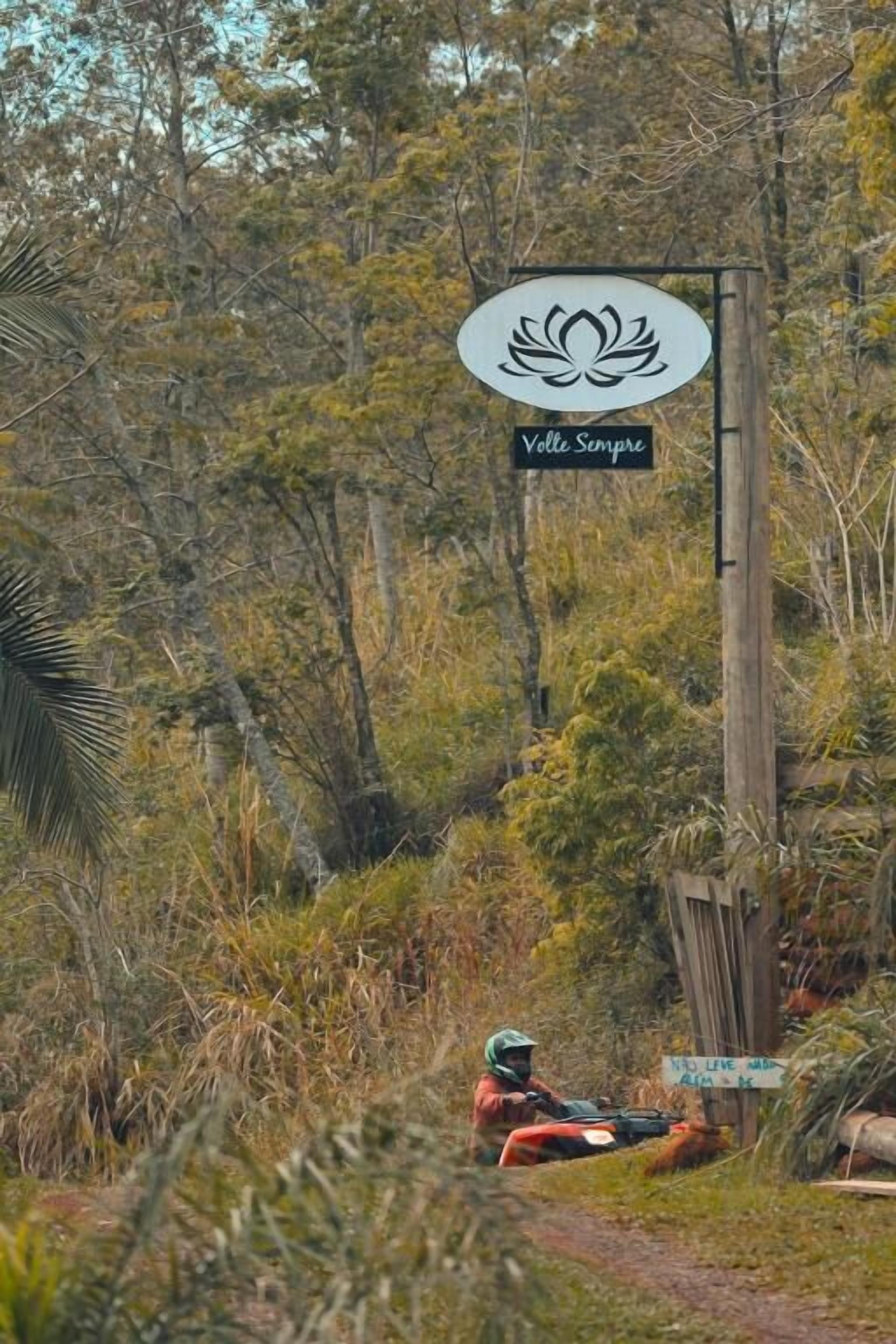 Une pancarte ornée d’un symbole de lotus et portant l’inscription « Velle Sempre », le tout dans un environnement naturel caractérisé par une végétation luxuriante.