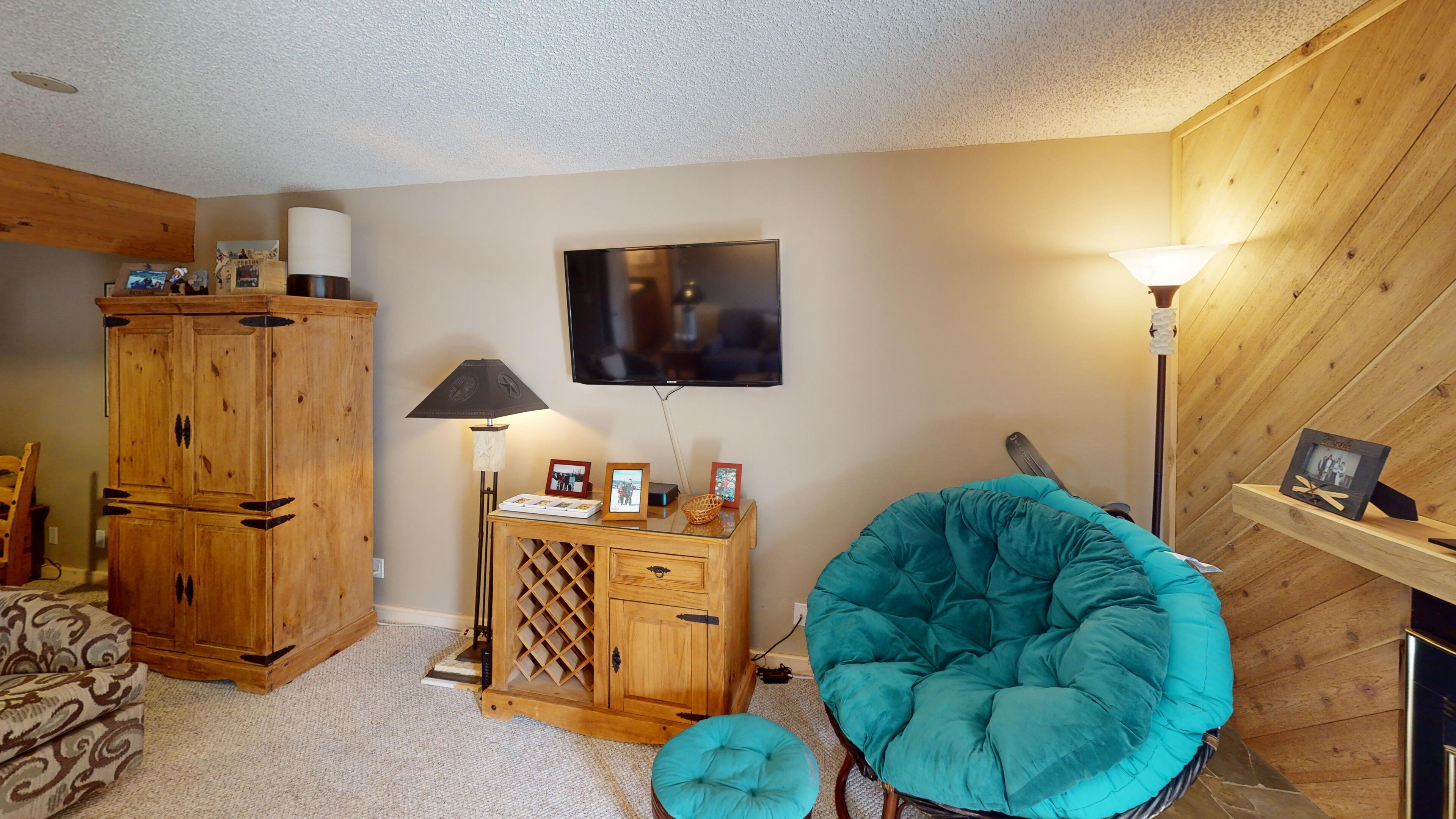 Un salon chaleureux avec un meuble en bois, un téléviseur fixé au mur, un fauteuil turquoise et une petite table en bois sur laquelle est posée une lampe.