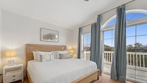 A bedroom with a large bed, a nightstand, a lamp, a framed picture on the wall, and a balcony with curtains.