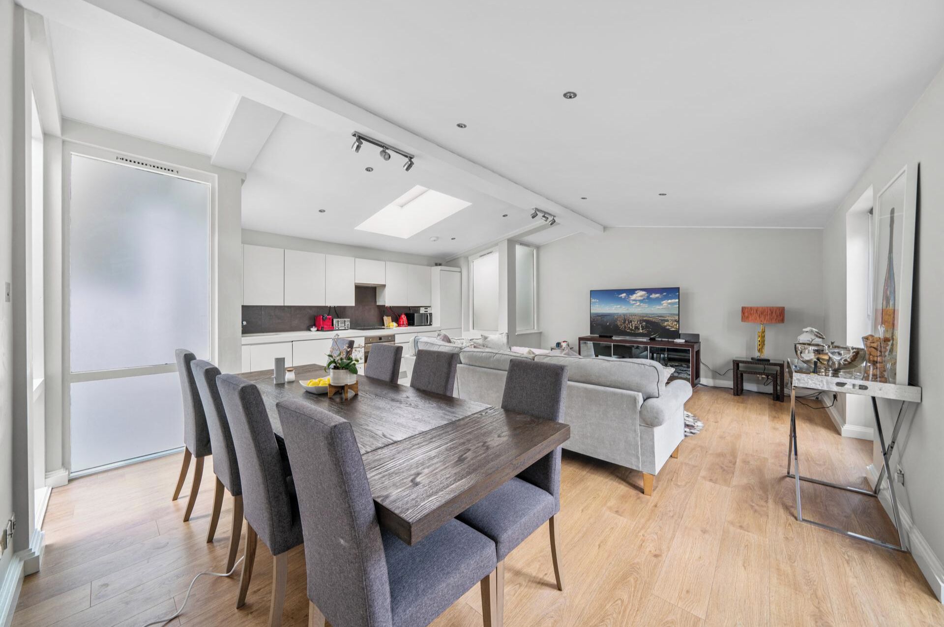 Una cocina moderna con armarios blancos, un comedor con mesa de madera y sillas tapizadas de gris, un sofá de color gris y un televisor en la sala de estar.