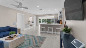 Un salon moderne avec un canapé bleu, une table basse en bois et un tapis à motifs. On y trouve également un coin cuisine avec un bar et des tabourets, ainsi qu’une grande fenêtre donnant sur un paysage verdoyant.