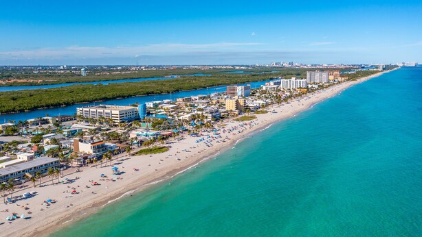 Vue aérienne d’une ville côtière avec une longue plage, des eaux d’un bleu limpide et un mélange de bâtiments résidentiels et commerciaux.