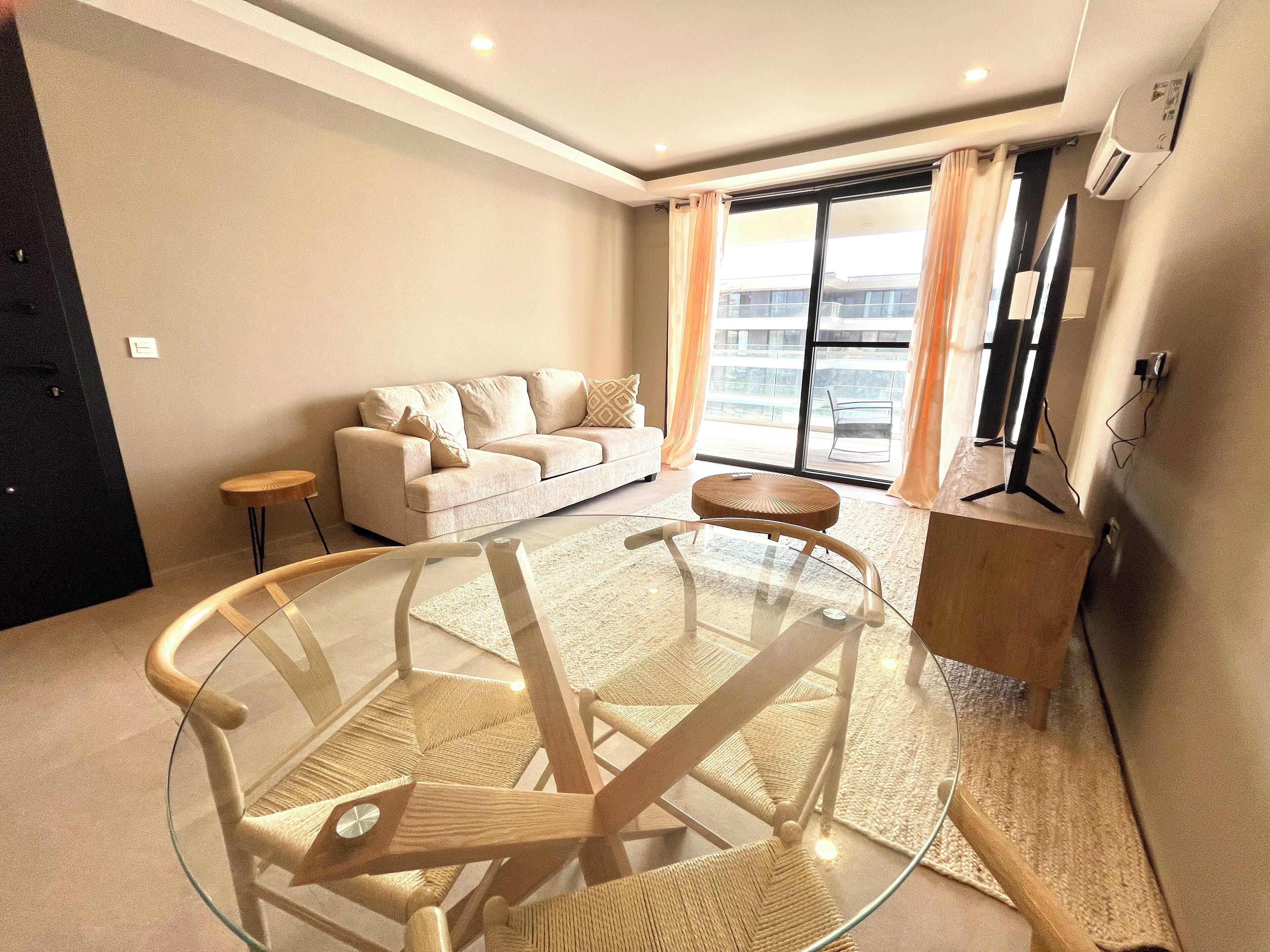 A modern living room with a glass-top coffee table, a beige sofa, and a view of the outside through large windows.