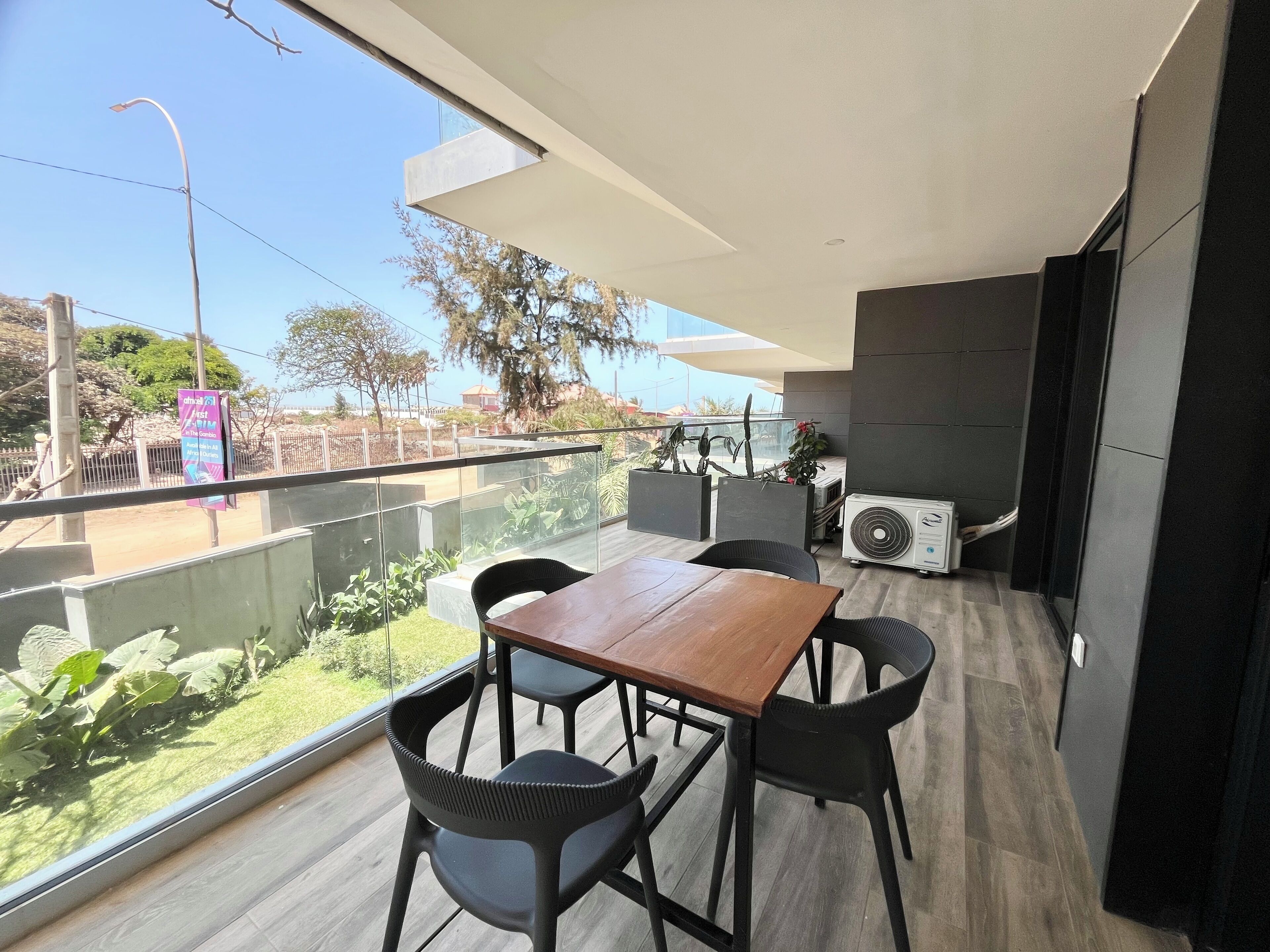 A balcony with a wooden table and chairs, a glass railing, and an air conditioning unit.