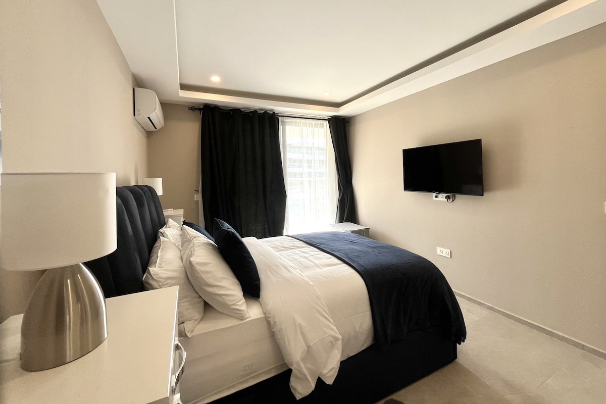 Una habitación de hotel moderna con una cama bien hecha, una mesita de noche con una lámpara, un televisor de pantalla plana en la pared y una ventana con cortinas.