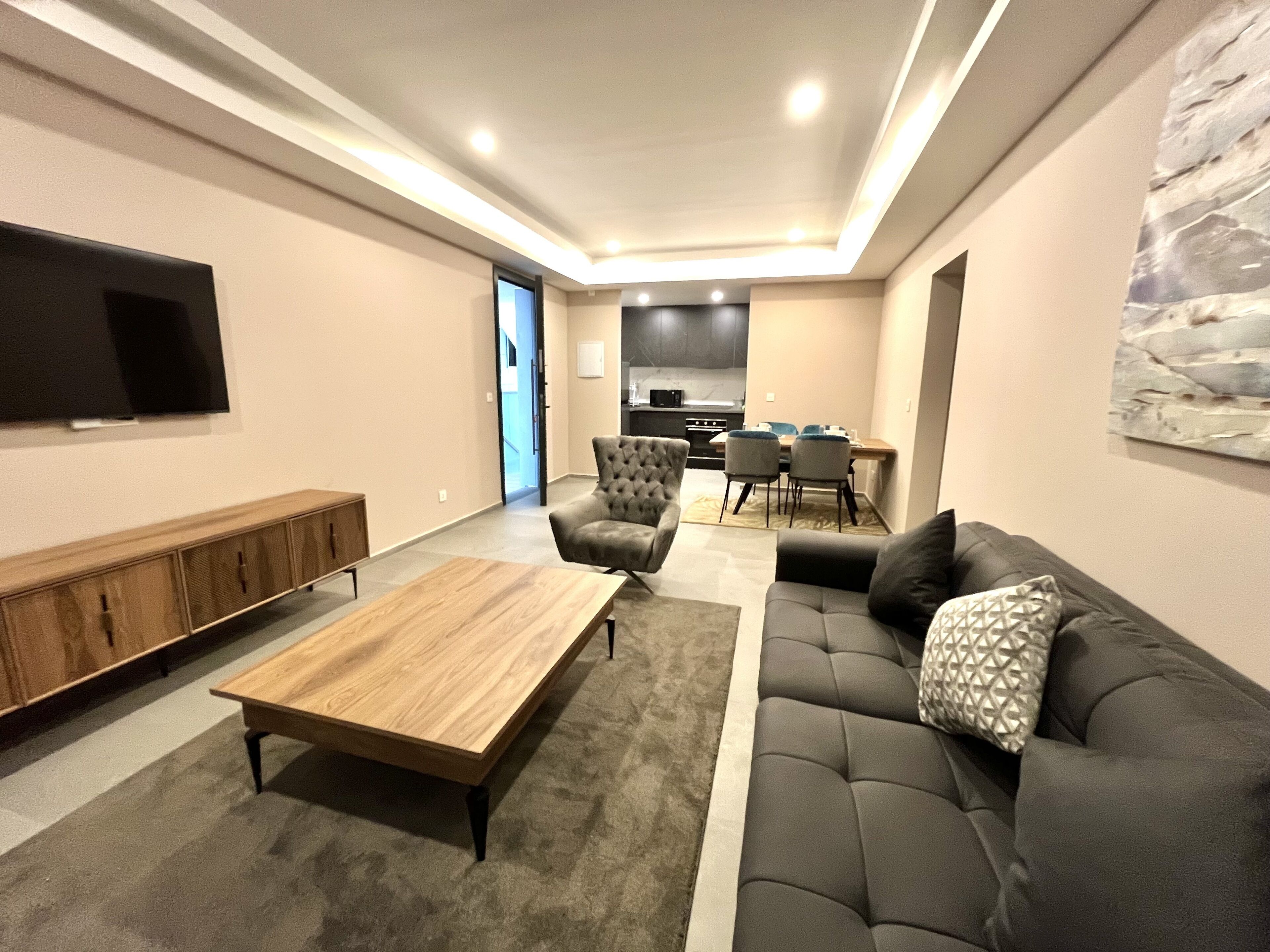 A modern living room with a sofa, coffee table, and TV. There is a dining area with chairs and a table in the background.