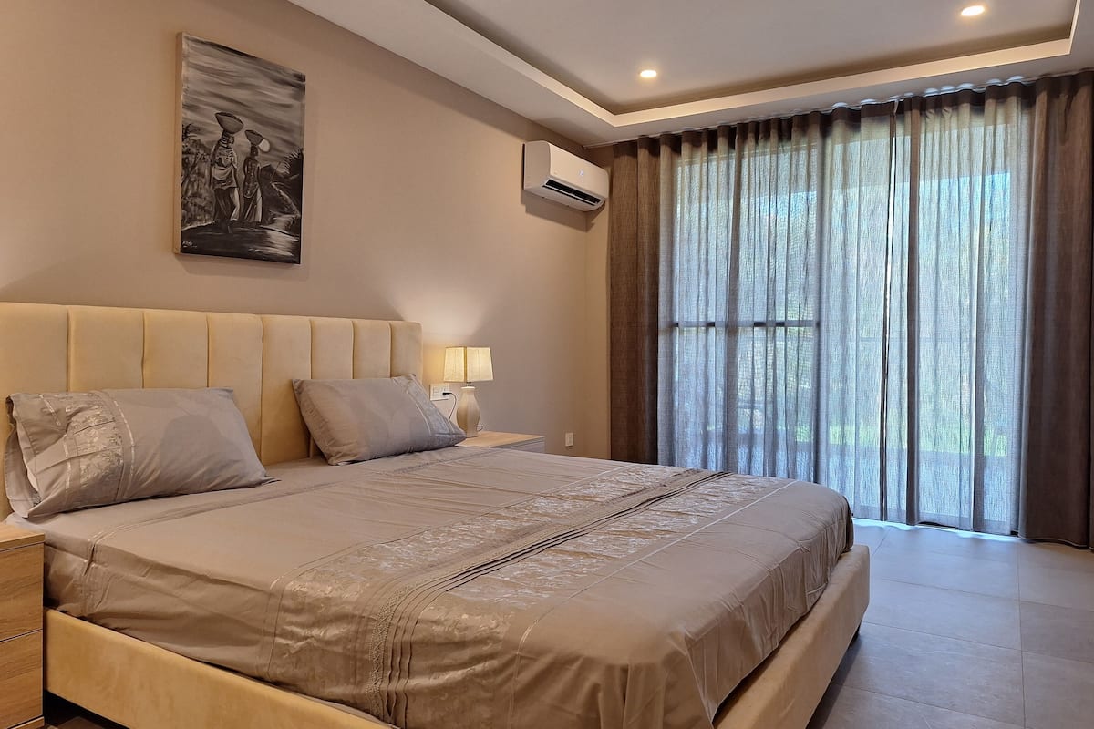 Un dormitorio con una cama grande, una mesita de noche con lámpara, un equipo de aire acondicionado y una ventana con cortinas.