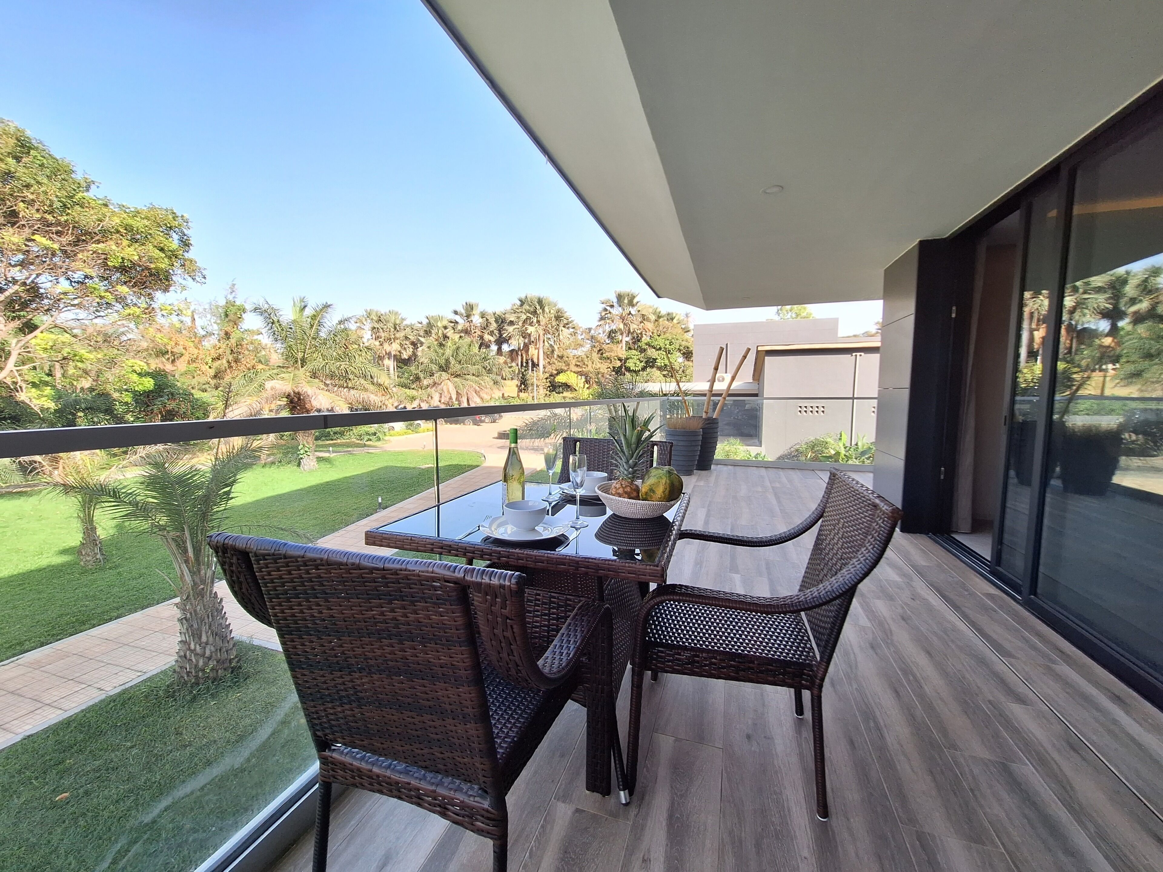 Un balcon avec une table dressée pour deux personnes, donnant sur un jardin agrémenté de palmiers et offrant une vue sur un ciel dégagé.