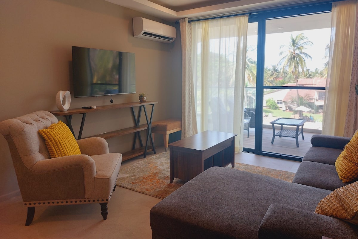 Una sala de estar con sofá, sillón y televisor sobre un mueble bajo. Hay una mesa y una silla cerca de la ventana, que ofrece vistas al exterior.
