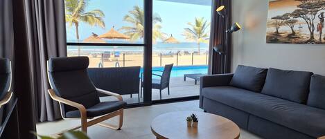 A modern hotel room with a balcony overlooking a beach, a sofa, a round table, and a painting on the wall.