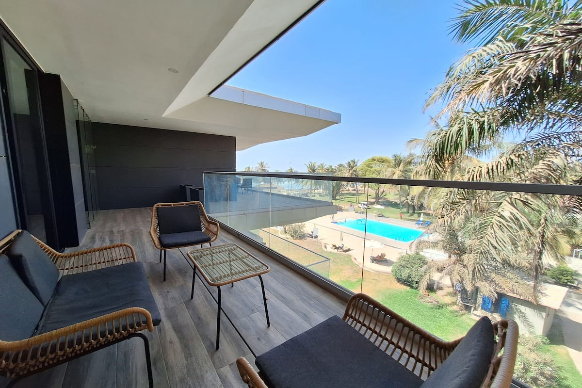 Un balcón con mobiliario de mimbre, baranda de vidrio y vista a una piscina y palmeras.