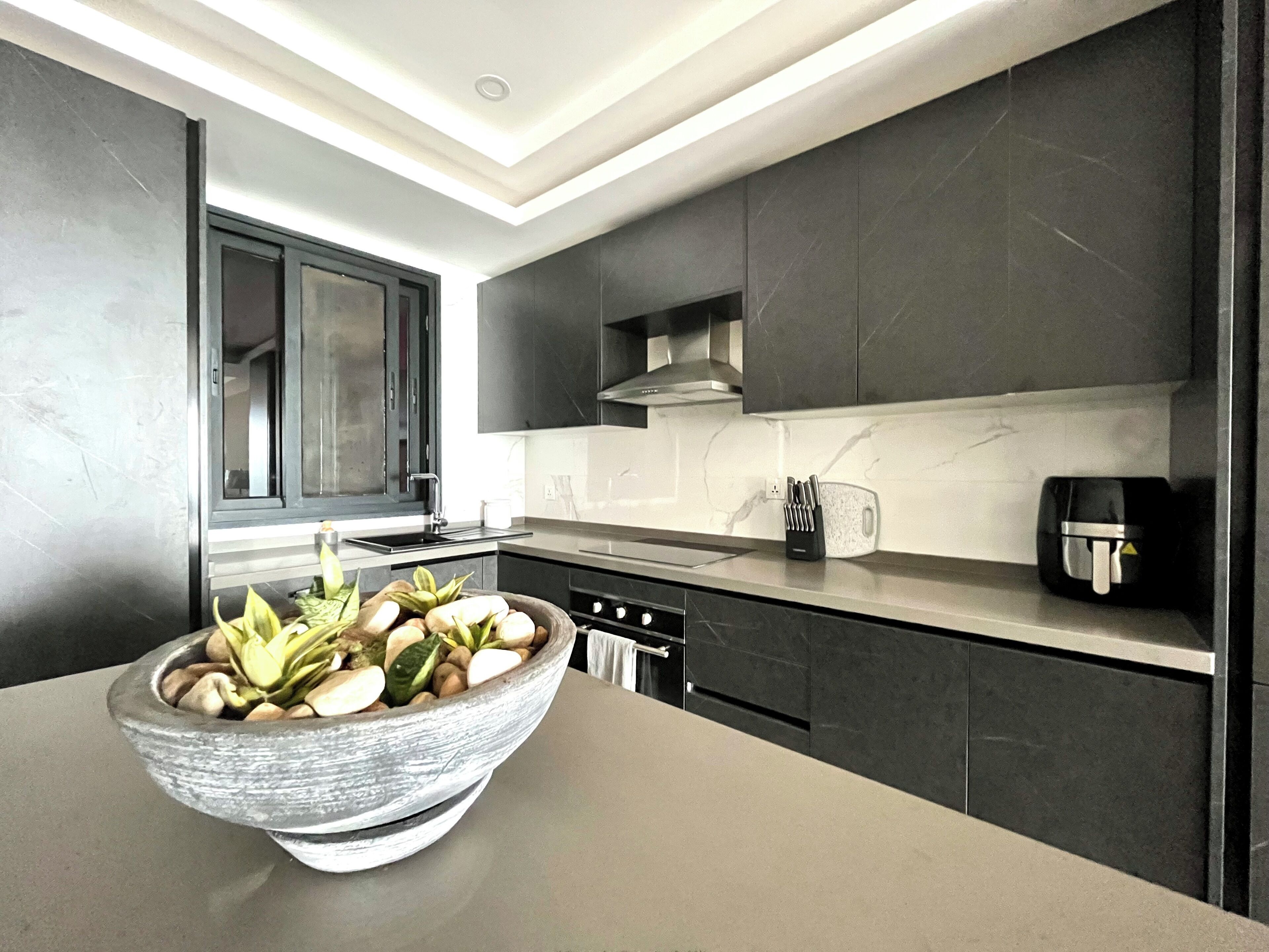 A modern kitchen with dark cabinetry, a large island with a bowl of fruit, and a coffee machine.