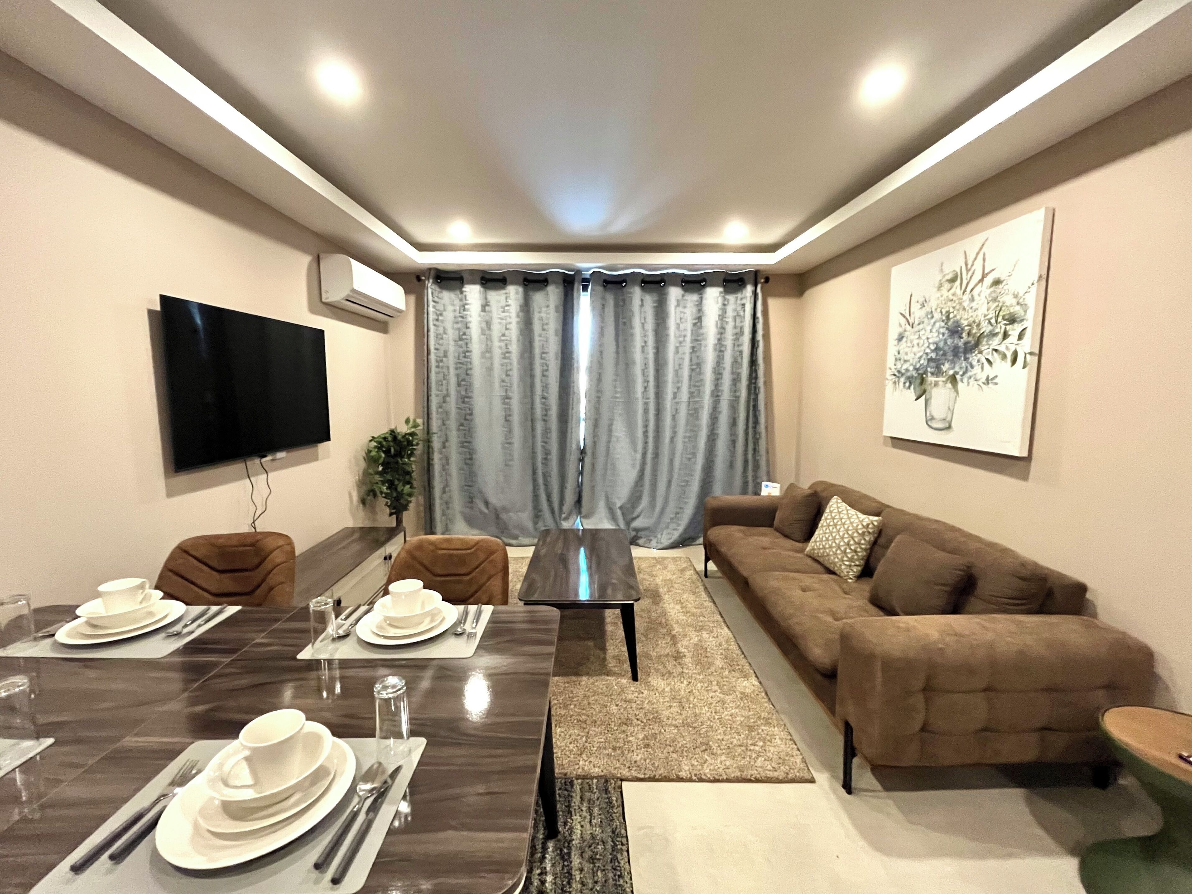 A modern living room with a dining table set for four, a brown sofa, a flat-screen TV mounted on the wall, and a large window with curtains.