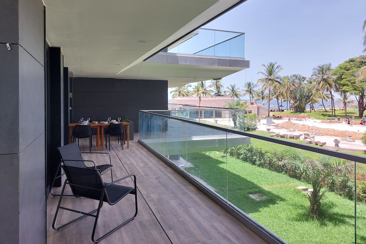 Un balcón moderno con barandilla de vidrio que da a un jardín ajardinado y una playa.