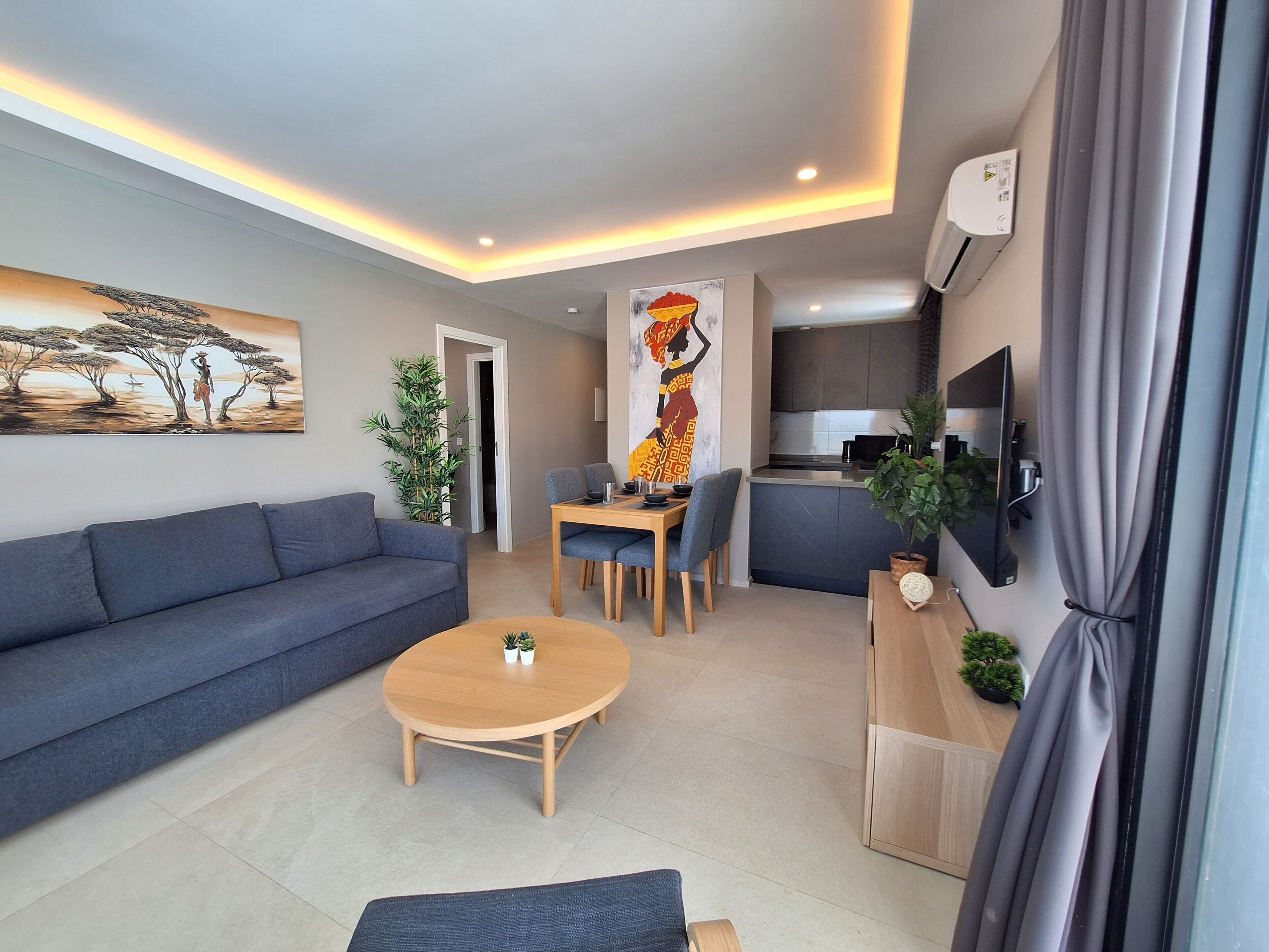 A modern living room with a sofa, a coffee table, and a dining area with chairs and a table. There is a kitchen area with a TV and a plant on the counter.