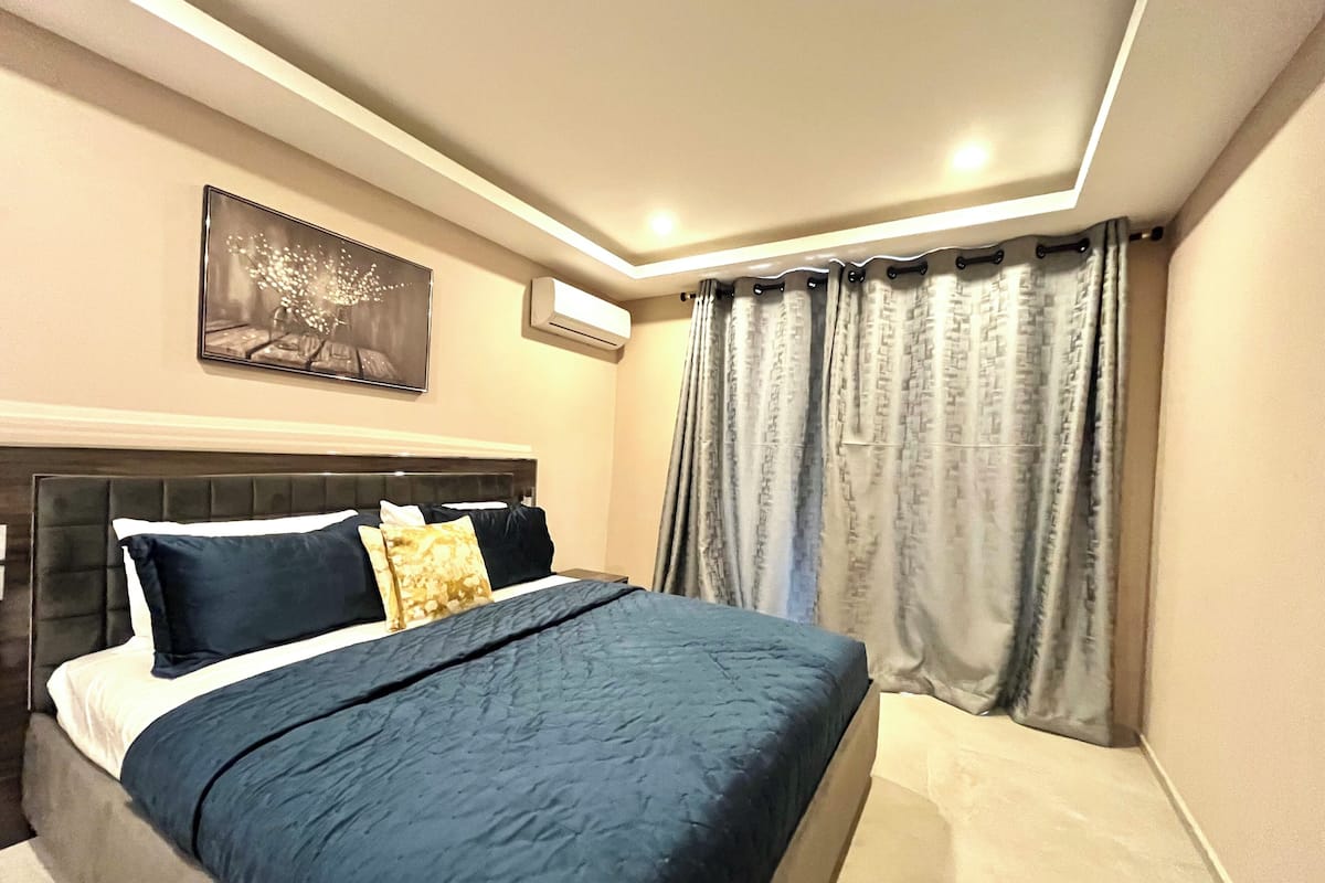 Un dormitorio moderno con una cama grande, cabecera, aire acondicionado de pared y ventana con cortinas estampadas.