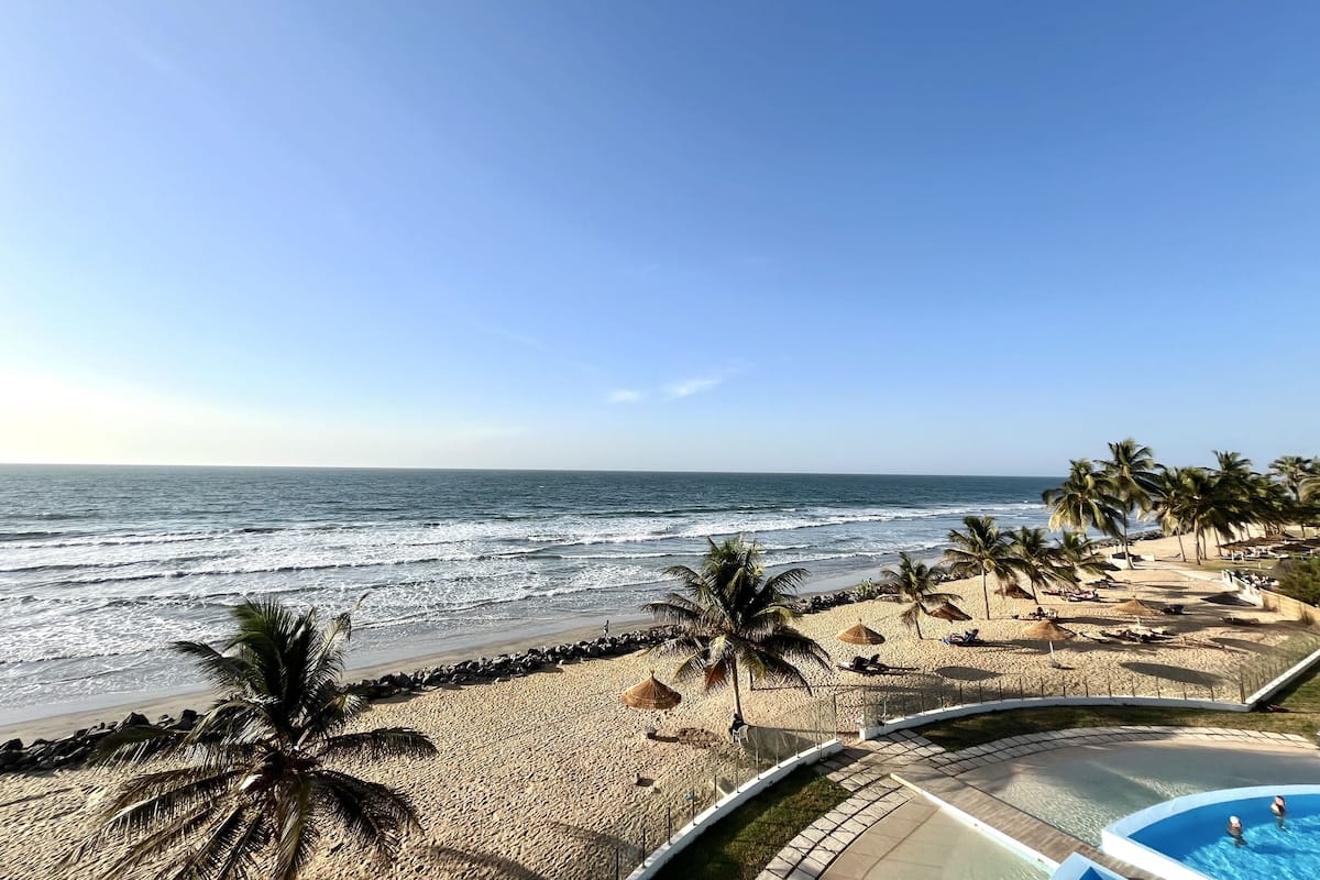 Vista a la playa con zona de piscina, palmeras y cielo despejado.