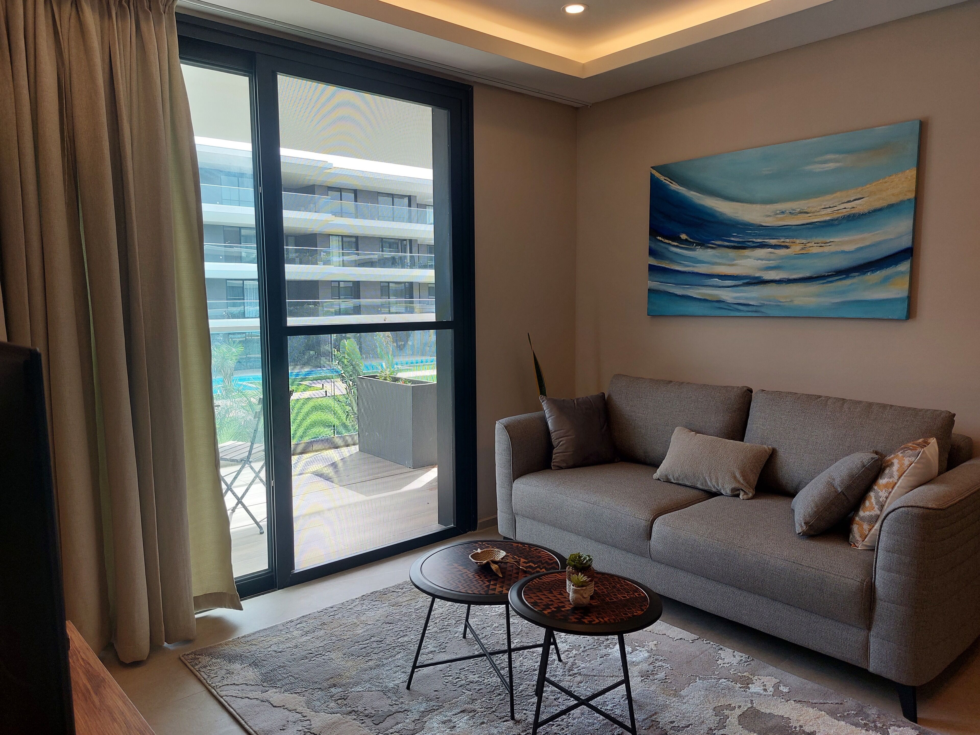 A modern living room with a grey sofa, a small round table, and a large window with a view of a building and greenery outside.