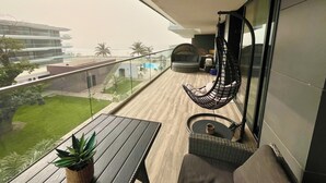 Un balcon avec un fauteuil suspendu, une petite table avec une plante, et une vue sur une piscine et des palmiers.